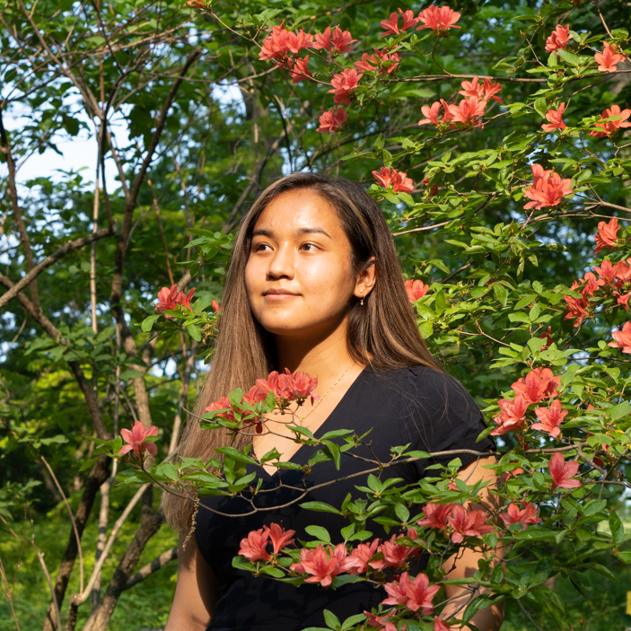 Kathleen Esfahany stands in the middle of a lush forest environment, framed by pink flowers. She is wearing a black dress and looking into the distance. 